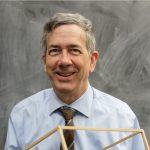 Photo of John Roeder in front of a chalkboard, wearing a blue shirt and tie.
