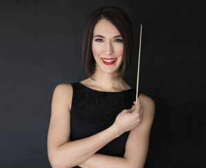Photo of Janna Sailor holding a baton in front of a dark background. She is wearing a black sleeveless dress.
